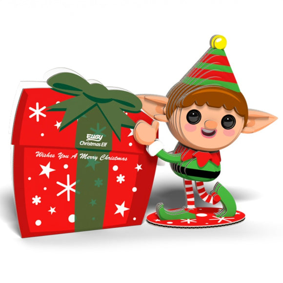 A charming Eugy Christmas Elf, from the EUGY brand, stands beside a large red and green eco-friendly gift decorated with white stars and snowflakes. The gift features a note that reads, "Wishes You a Merry Christmas." The elf sports a playful striped hat and green outfit, reminiscent of the whimsical Eugy toy figures.