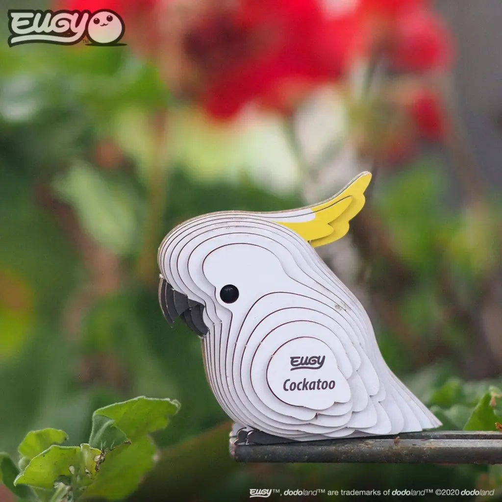 A decorative and eco-friendly 3D puzzle cockatoo with layered white pieces and a yellow crest perches on a railing, surrounded by green leaves and a flower. "Eugy Cockatoo" by EUGY is printed on its side for an engaging model-building experience.