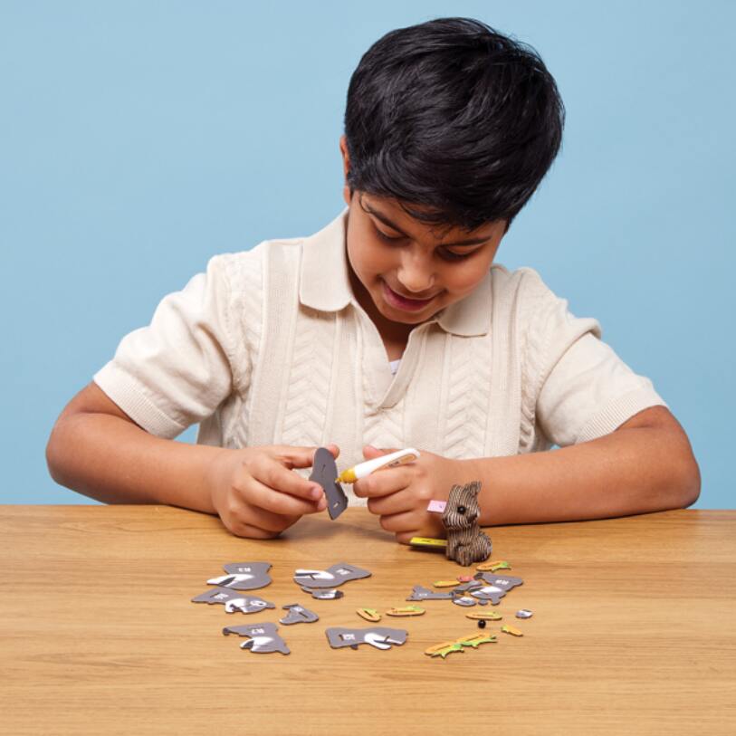 A young boy sits at a wooden table against a blue background, concentrating on assembling pieces of the EUGY Raccoon puzzle by EUGY. The pieces are shaped like various animals, resembling miniature 3D models. He carefully fits two pieces together while a small toy raccoon sits nearby.