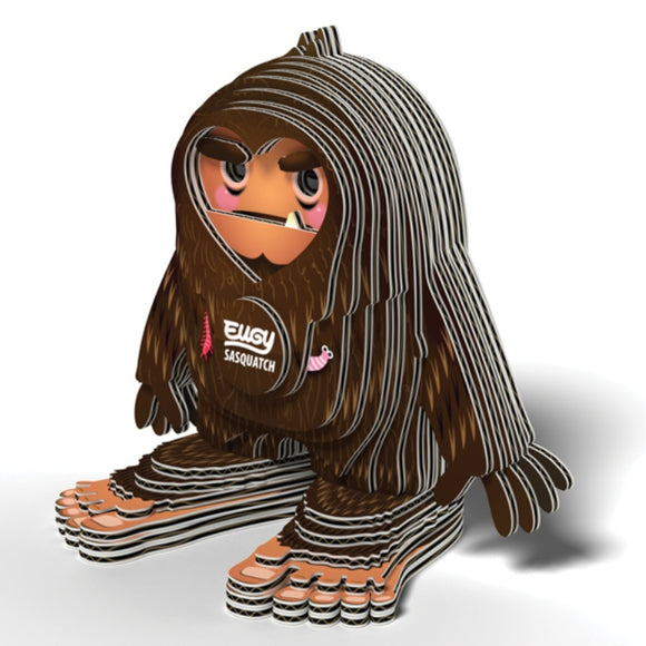 Introducing the EUGY Sasquatch Big Foot by EUGY: a whimsical, layered cardboard cutout of a creature akin to Bigfoot. Crafted from eco-friendly materials, it boasts large feet, floppy arms, and a shaggy brown appearance. The front is adorned with the playful and stylized "Sasquatch" and "Ellgy" design.