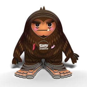 The EUGY Sasquatch Big Foot by EUGY is a stylized, cartoonish figure featuring brown fur, pink cheeks, and wide feet made from sustainable materials. It has a stern expression with small decorative details such as a pink worm and feather. Across its chest, "Elusive Sasquatch" is printed in white text.