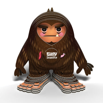 The EUGY Sasquatch Big Foot by EUGY is a stylized, cartoonish figure featuring brown fur, pink cheeks, and wide feet made from sustainable materials. It has a stern expression with small decorative details such as a pink worm and feather. Across its chest, "Elusive Sasquatch" is printed in white text.