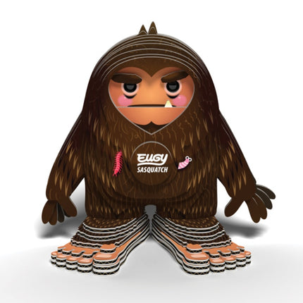 The EUGY Sasquatch Big Foot by EUGY is a stylized, cartoonish figure featuring brown fur, pink cheeks, and wide feet made from sustainable materials. It has a stern expression with small decorative details such as a pink worm and feather. Across its chest, "Elusive Sasquatch" is printed in white text.