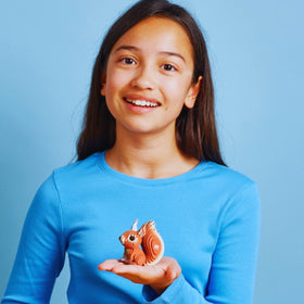 A young girl in a blue shirt smiles as she holds the EUGY Squirrel, an eco-friendly figure by EUGY, in her open palm against a light blue background.