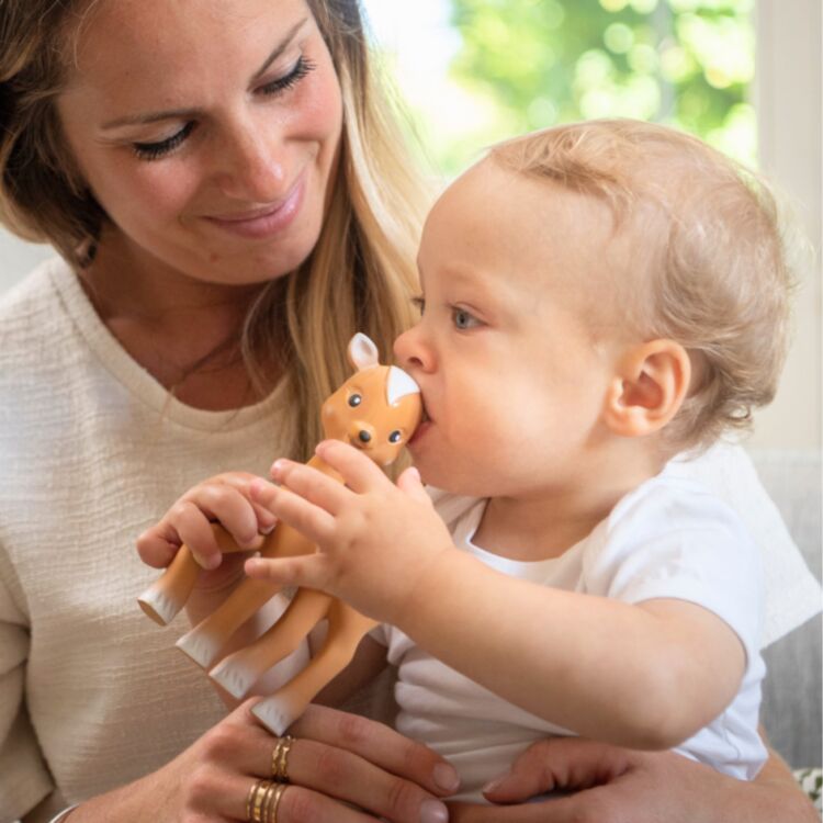 A woman with long hair smiles warmly while holding a baby, who is biting Fanfan the Fawn from Sophie La Giraffe. The baby, dressed in a white shirt, sits contentedly on the sofa with a bright window in the background.