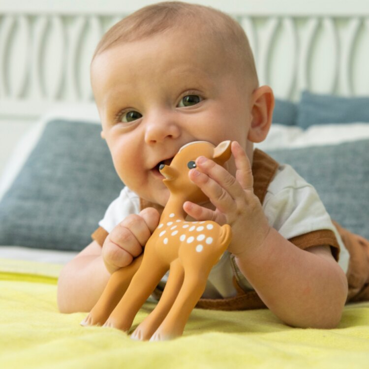 A baby, dressed in a white shirt and brown overalls, smiles while lying on a yellow blanket holding Fanfan the Fawn by Sophie La Giraffe. Nearby is a natural rubber teether, set against a backdrop of a white headboard and blue pillows.