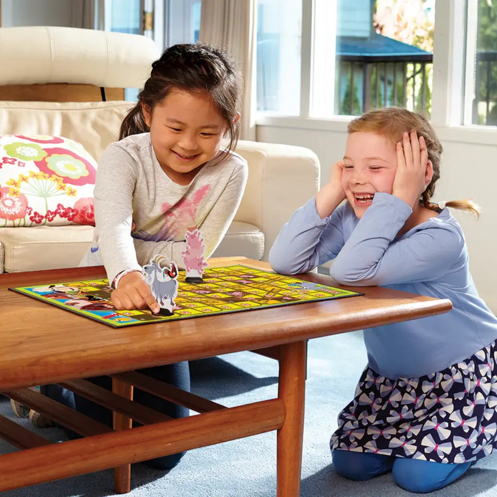 Two young girls play Farm Snakes & Ladders by Outset Cheatwell on a wooden table. The girl in the gray shirt moves a piece while the one in the blue top smiles, hands on cheeks. A couch and bright window complete this delightful family board game scene.