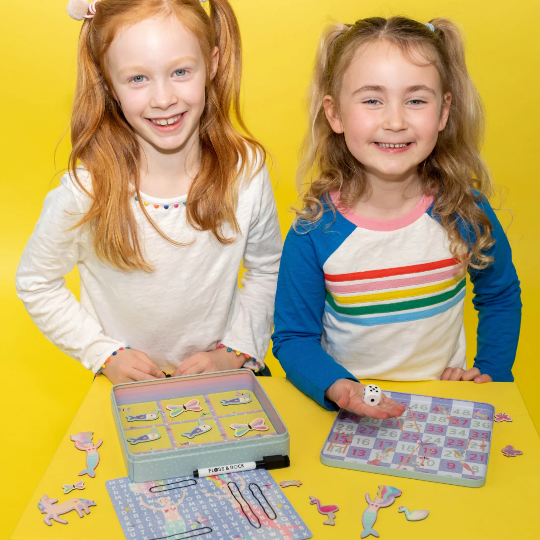 Two young girls smile at the camera while playing with the eco-friendly Fantasy Magnetic Fun & Games by Floss & Rock. One holds a die, and the other is near a game featuring animal pieces made from recycled materials. Both are standing against a bright yellow background.