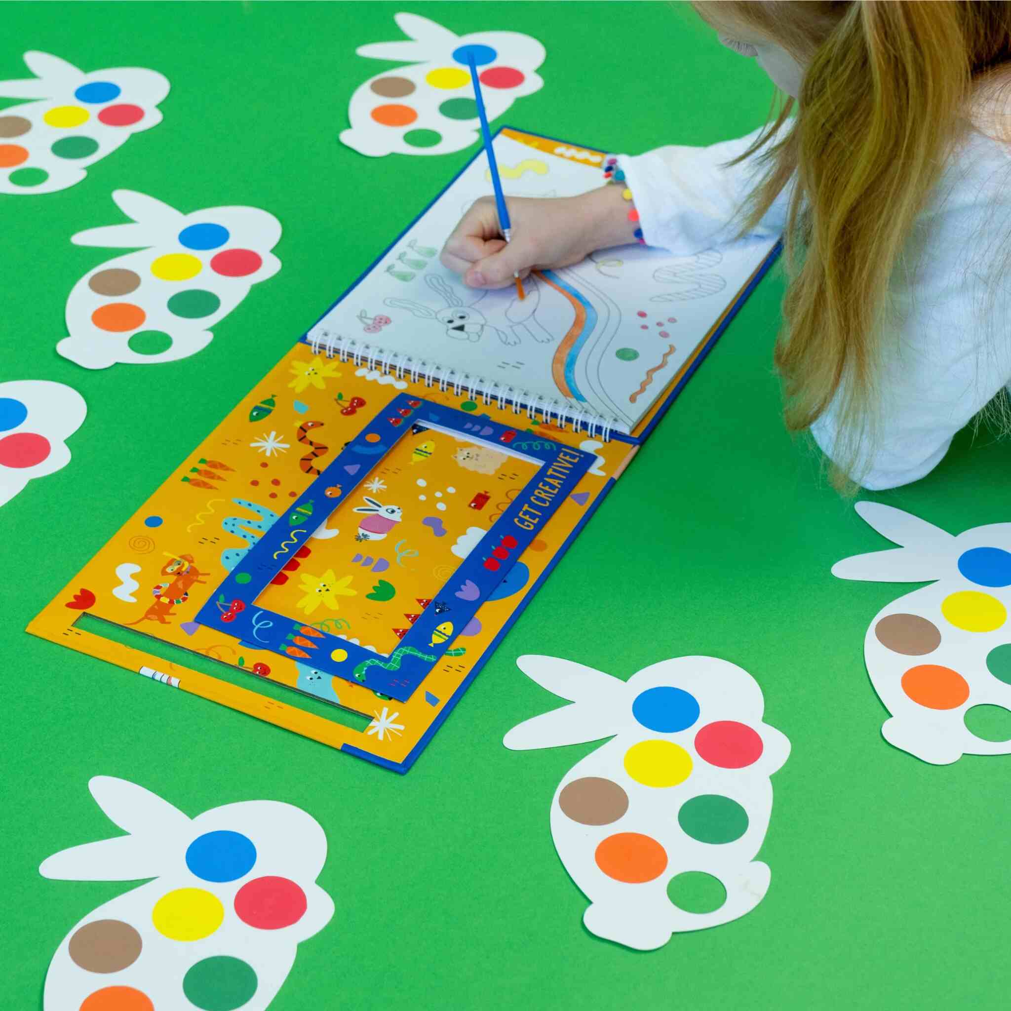 A child drawing on the vibrant Floss & Rock Pets My Painting Pad, placed on a green surface, with rabbit-shaped cutouts adorned with multicolored dots. The child, wearing a white shirt and using a blue pencil, is inspired by the playful design of these eco-friendly rabbit-shaped palettes scattered around.