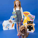 A young girl with red hair stands smiling on a blue background, holding a book. She wears overalls and is next to a curly-haired dog on a leash. Behind them are colorful cartoon cutouts of animals from the "Pets Magnetic Playtime" collection by Floss & Rock, cleverly crafted from recycled materials with playful expressions.