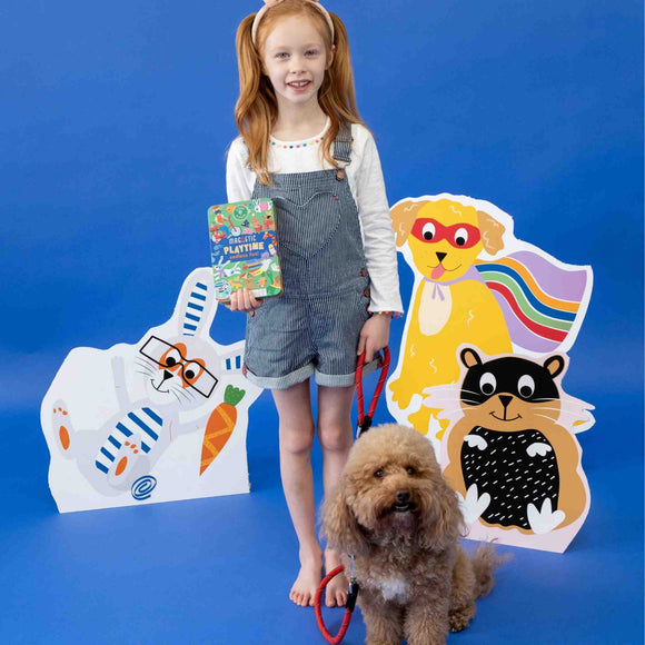 A young girl with red hair stands smiling on a blue background, holding a book. She wears overalls and is next to a curly-haired dog on a leash. Behind them are colorful cartoon cutouts of animals from the "Pets Magnetic Playtime" collection by Floss & Rock, cleverly crafted from recycled materials with playful expressions.