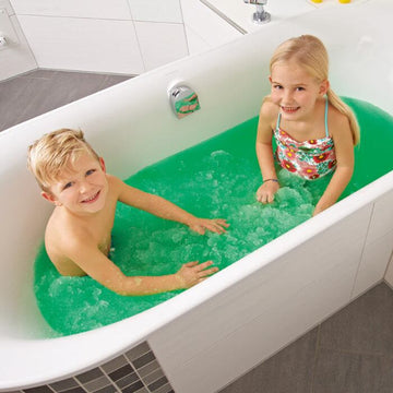 Two children are sitting in a bathtub filled with Zimpli Kids' Gelli Baff. The boy is on the left, and the girl on the right wears a colorful swimsuit. They are smiling and playing happily in the bathroom, which has white tiles and a grey floor.