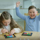 A boy and a girl play The Genius Square by The Happy Puzzle Company at a table. The boy, in a blue shirt, smiles widely with raised arms after rolling the dice, while the girl with braided hair focuses on arranging blocks.