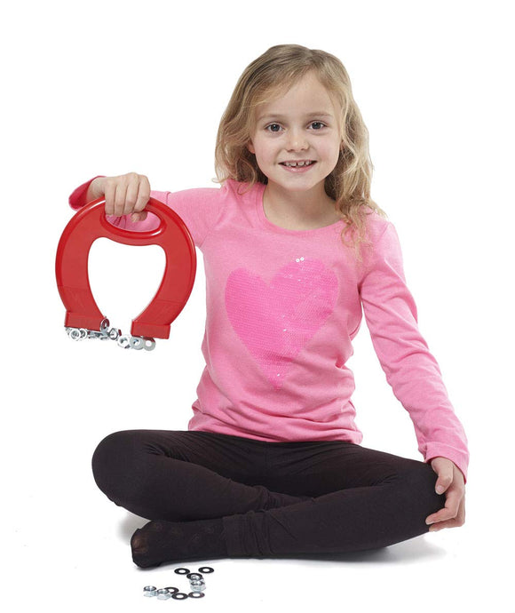 A child in a pink shirt and black pants sits cross-legged on the floor, holding a Massive Magnet from BRAINSTORM TOYS. It's one of her favorite STEM toys, attracting small metal objects. She smiles at the camera, captivated by the wonders of magnetism.