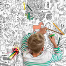 A child with blond hair and a striped shirt colors with markers on an eggnogg Giant Poster/Tablecloth – Countryside, featuring outlined illustrations of plants, animals, and shapes. A horse is partially colored in orange and green, while colored pencils nearby await more fun coloring activities.