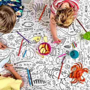 Three children color an eggnogg Giant Poster / Tablecloth – Seaside, featuring sea creatures and designs. Crayons, colored pencils, apple slices, and drinks scattered around create a lively scene perfect for children's parties.