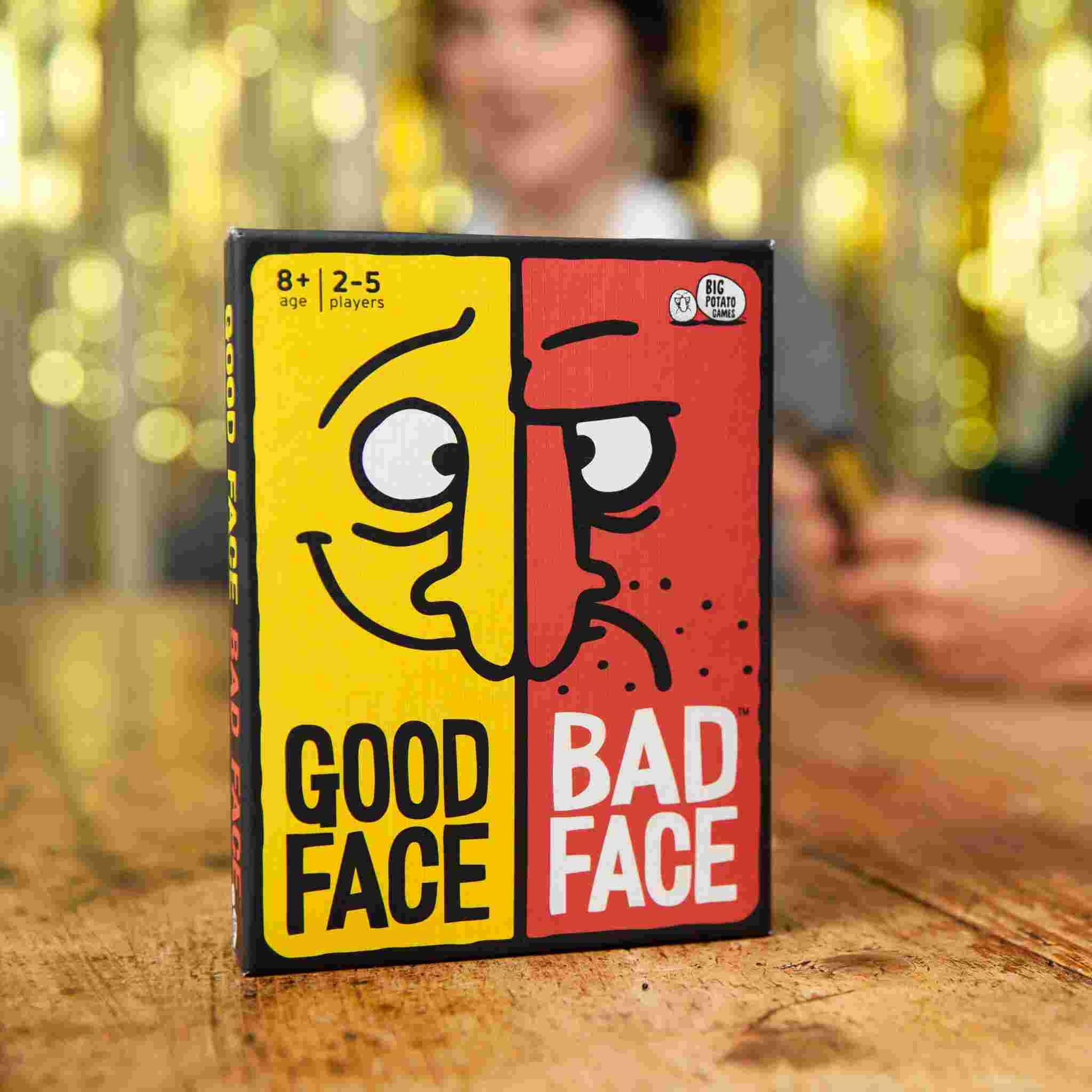 A board game box from Big Potato, titled "Good Face Bad Face," rests on a wooden table. The design features two distinctive halves—one yellow with a smiling cartoon face and one red with a frowning cartoon face. This fast-paced party game is ideal for 2-5 players, ages 8 and up.