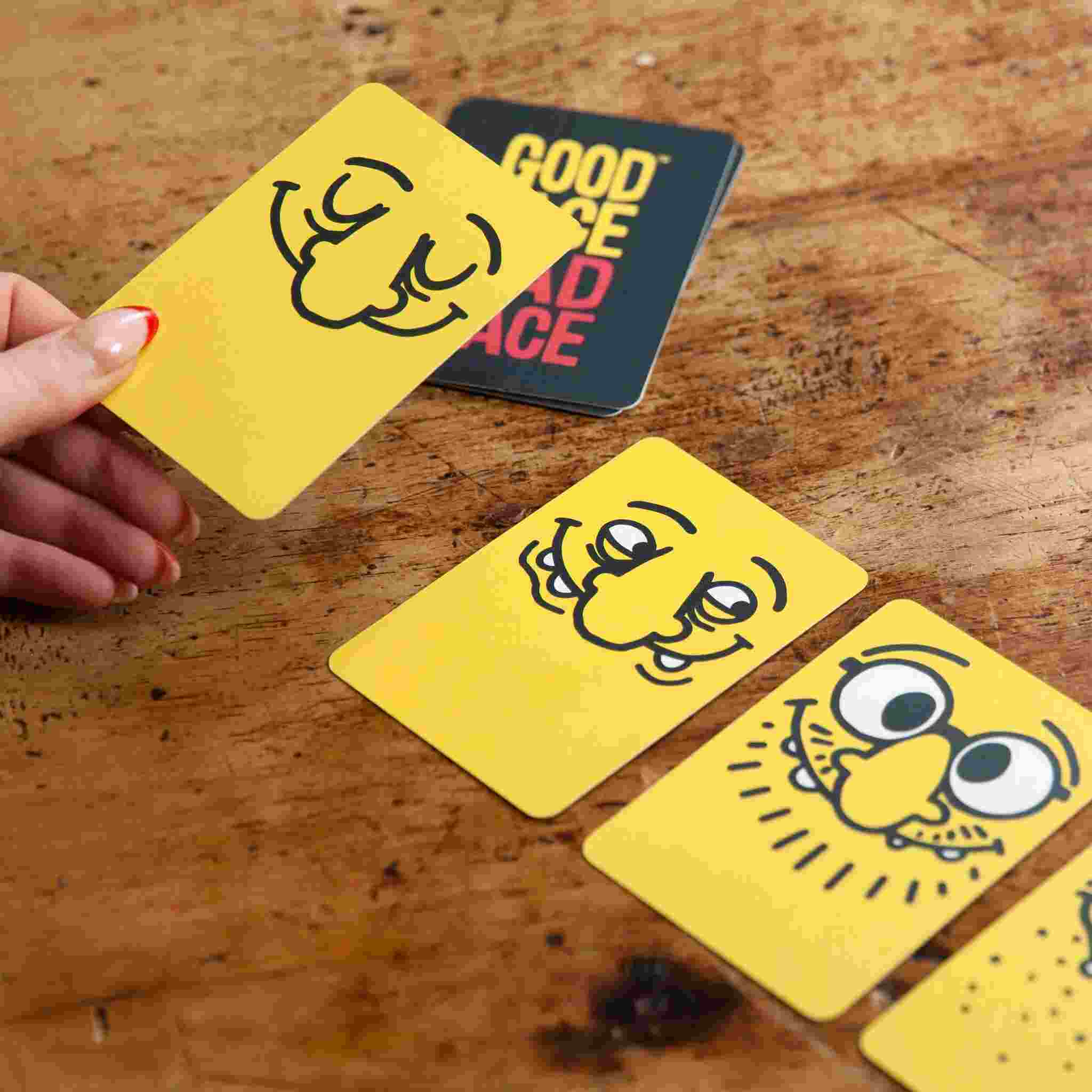 A hand holds a yellow card from Good Face Bad Face by Big Potato, featuring a cheerful face drawing, ideal for an exciting card game. Three similar cards are laid out on the wooden surface, suggesting the enjoyment of this party game. In the background, a black card with red and yellow text is partially visible.