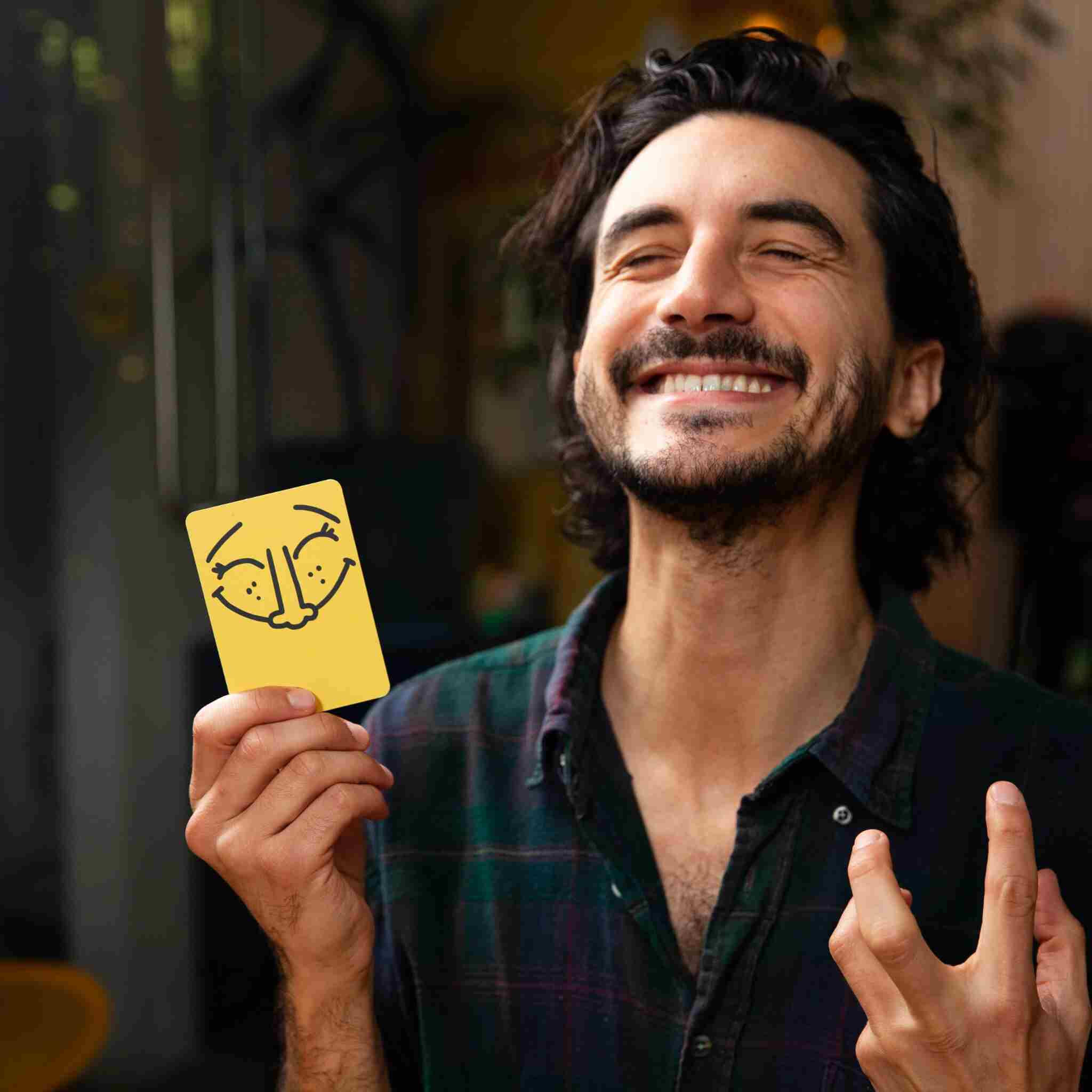 A person with a broad smile holds a card from "Good Face Bad Face" by Big Potato, featuring a humorous face drawing. They're wearing a dark plaid shirt and appear joyful in the cozy indoor setting, as if enjoying a fast-paced party game.