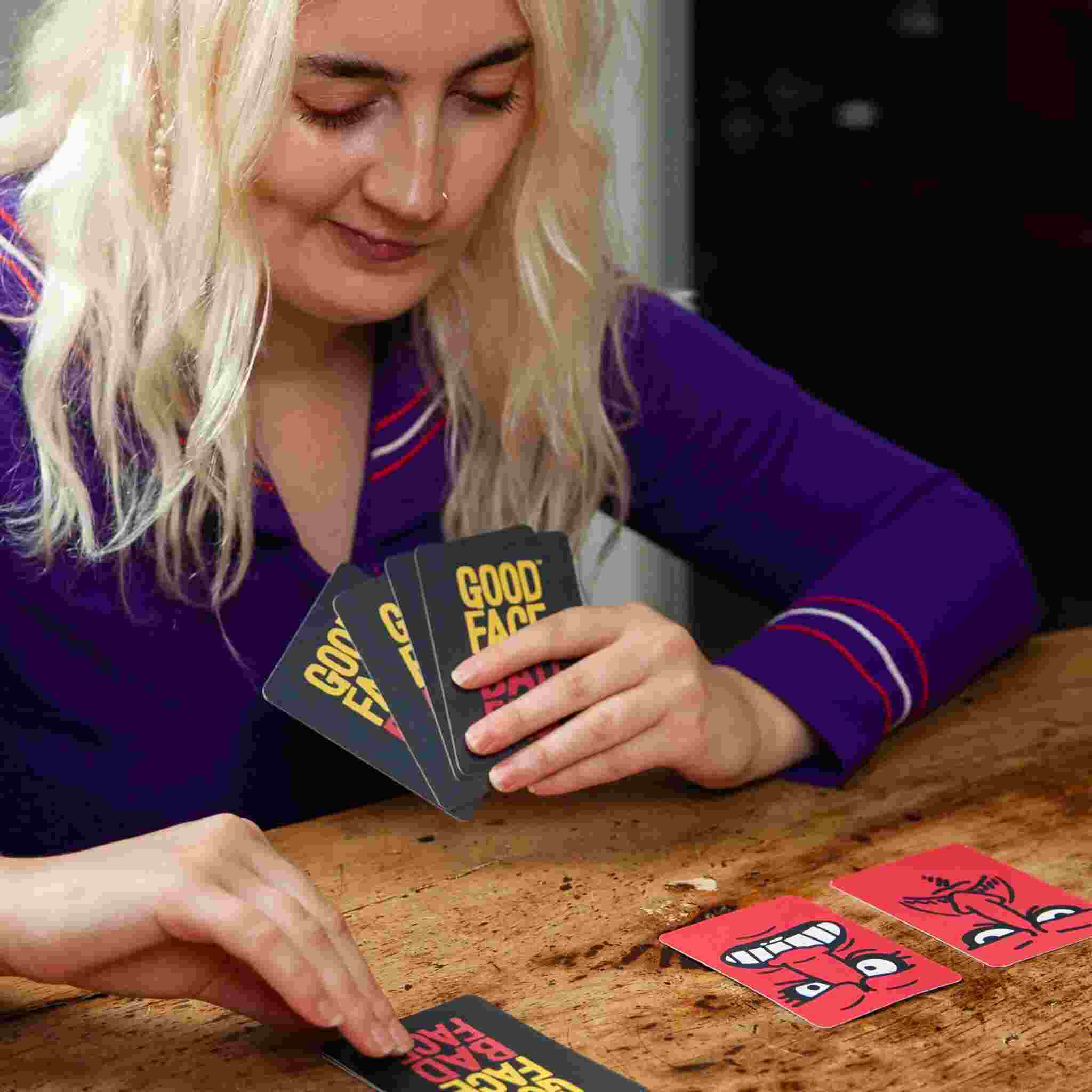 A person with long blonde hair is deeply engaged in a fast-paced card game at a wooden table. They are holding cards from "Good Face Bad Face" by Big Potato and placing one on the table, where lively face illustrations enhance the excitement of this party game.