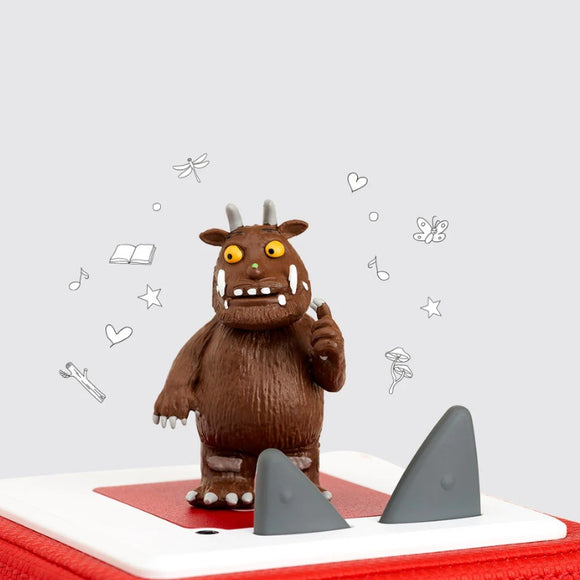 A small figurine of the Gruffalo, from Tonies' "Tonie - The Gruffalo" collection, inspired by Julia Donaldson's tale, features horns, sharp teeth, and a raised finger as it stands on a red base. Surrounding it are icons like books, musical notes, hearts, swimming fish, and stars set against a light grey background.