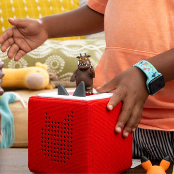 A child with brown skin, wearing an orange shirt and a turquoise watch, stands beside a red Tonies speaker featuring the Gruffalo figurine from the Tonie - The Gruffalo collection. In the background, there is a sofa and a cushion with various patterns, as well as a toy yellow dog, evoking the charm of Julia Donaldson's tales from the deep dark wood.