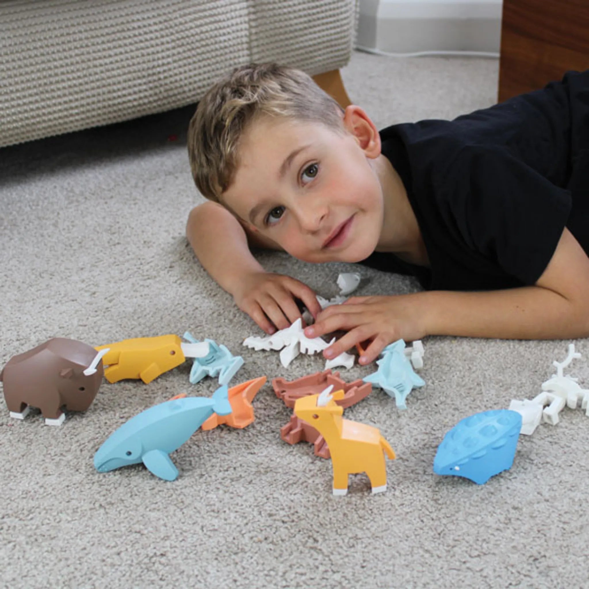 A young boy smiles at the camera while lying on a carpet, surrounded by colorful Halftoys World set – Animal creatures, including a whale, bison, dinosaur, and more from the Halftoys brand.