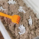 Play sand with white dinosaur skeleton toys and an orange rake in a container