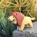 The Halftoys Diorama - Lion by Halftoys is a small, blocky lion toy with a brown mane and cartoon-like design, perfect for animal-themed playsets or a lion diorama. It stands on concrete next to green spiky plants.