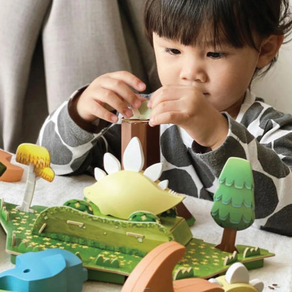 A young child in a patterned shirt carefully plays with the Halftoys Diorama - Stego by Halftoys, an educational dinosaur toy and tree puzzle set that encourages interactive STEM learning.