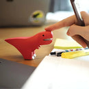A hand touches the nose of a Halftoys Diorama - T-Rex by Halftoys, a small red educational dinosaur toy on a desk beside pens, a highlighter, and an open notebook, with a laptop and sticky notes in the background.