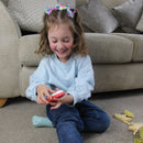 A young girl with curly hair and rainbow bows smiles as she sits cross-legged on a carpet, playing with the Halftoys Diorama - T-Rex by Halftoys. Wooden toys are scattered nearby, and a beige sofa is in the background.