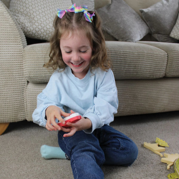 A young girl with curly hair and rainbow bows smiles as she sits cross-legged on a carpet, playing with the Halftoys Diorama - T-Rex by Halftoys. Wooden toys are scattered nearby, and a beige sofa is in the background.