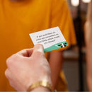 A person holding a small card with the question: "If you could have an extra eye somewhere on your body, where would you choose?" This intriguing question is featured in the party game Herd Mentality by Big Potato. In the blurred background, another person wearing a yellow shirt can be seen.