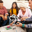 Four friends gather around a table, happily immersed in "Herd Mentality," the ideal party game by Big Potato. One of them holds the game box adorned with its distinctive cow pattern. The table is strewn with papers and a small pink cow toy, all set against the backdrop of a cozy living room.