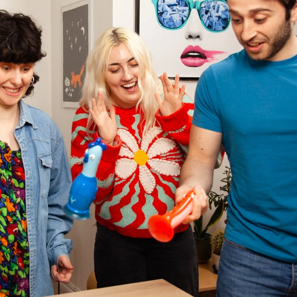 3 adults playing the Chicken vs Hot Dog game.  Blue chicken piece is in the air, lady in middle in red top laughing, man on right wearing a blue t-shirt is about to throw the hot dog piece.