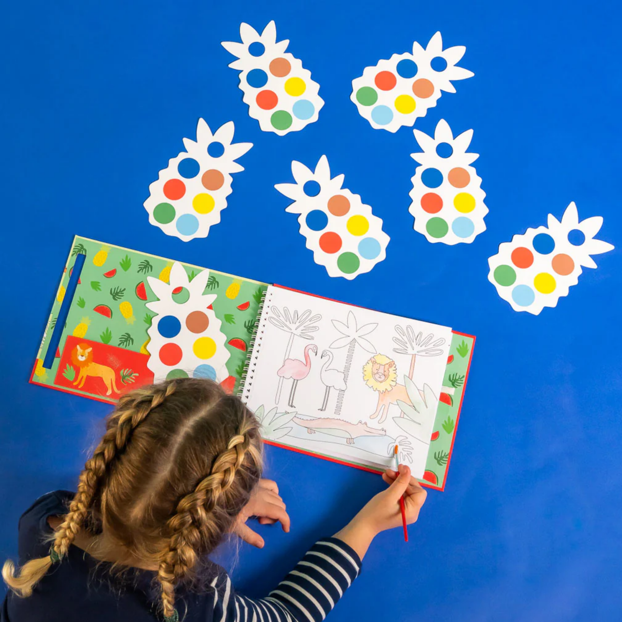 A young artist with braided hair colors in the Jungle My Painting Pad by Floss & Rock, which features a lion and flamingos. Nearby, pineapple-shaped paint palettes adorned with colorful circles lie on a blue surface. The child, dressed in a striped shirt, creatively engages in an eco-friendly activity.