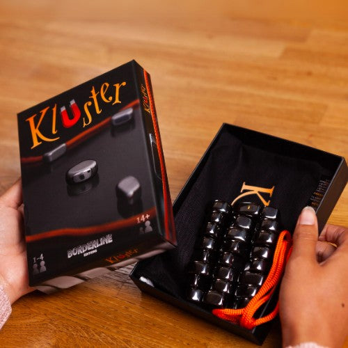 Hands holding a Kluster board game box and its contents by Big Potato Games. The open box reveals a pouch of black magnetic stones, ideal for this engaging magnetic party game, along with an orange string on a wooden table. The box features the Big Potato Games logo and recommends the game for ages 14 and up.