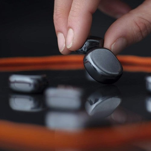 A hand with neatly manicured nails places a smooth, shiny black Kluster stone by Big Potato Games on a reflective surface. More stones are scattered around, with an orange line faintly visible in the background, reminiscent of pieces in a sophisticated game of Kluster.