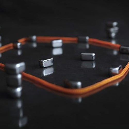 A captivating arrangement features a circular configuration of vertical, rectangular gray magnets on a glossy black surface, encircled by two intertwining, thin orange cords. This fascinating setup mimics the "Kluster" magnetic game by Big Potato Games, where magnets are playfully scattered both within and beyond the circle.