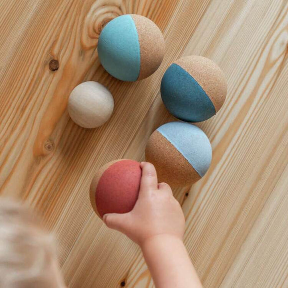 A child eagerly reaches for vibrant two-toned balls on a wooden surface from the Korko Champions Cork Boccia Set in teal, red, and beige, enhancing their fine motor skills.