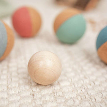 A close-up of a wooden ball on a textured surface, with blurred multicolored balls in the background, resembles the Champions Cork Boccia Set by Korko. Shades of pink, green, blue, and light brown evoke a playful and natural vibe that encourages social skill development.
