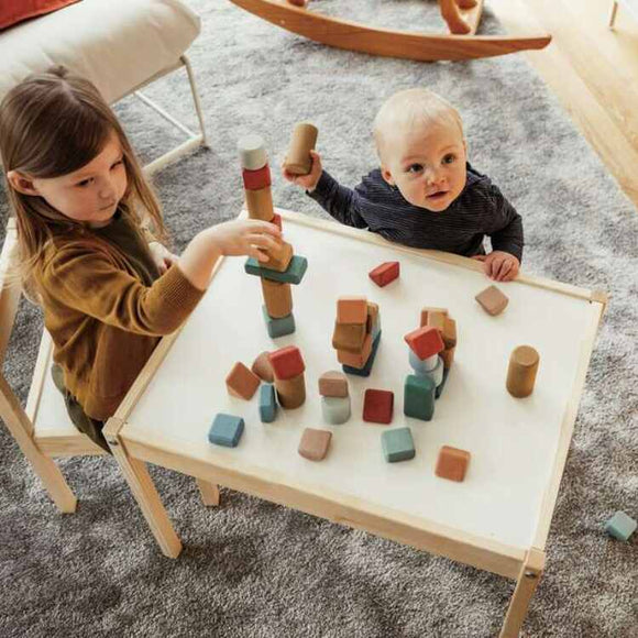 Two children happily play with eco-friendly toys at a small table. The girl stacks Korko's Giant Architects, 60 Cork Building Blocks to enhance her motor skills as the boy observes. A carpeted floor and a rocking horse are visible in the background.