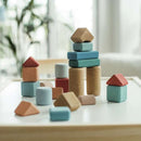 The table is adorned with Korko's Small Architects, 20 Cork Building Blocks in vibrant blue, red, and beige cubes, cylinders, and triangular prisms. These educational toys boost fine motor skills. A charming indoor setting with plants can be seen in the blurred background.
