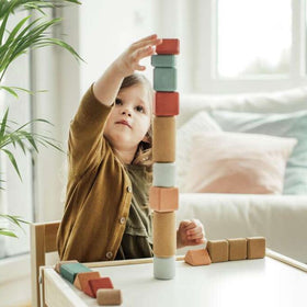 A young girl, focused intently, stacks Korko's Small Architects 20 Cork Building Blocks on a table, enhancing her fine motor skills. A plant and cushions in the background create a cozy setting.