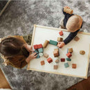 Two children play with the vibrant Korko Tall Architects, 40 Cork Building Blocks on a white table. One is light-haired, the other dark-haired, as they sit on a gray textured carpet, engaging in playful and educational activities that enhance fine motor skills through imaginative interaction.