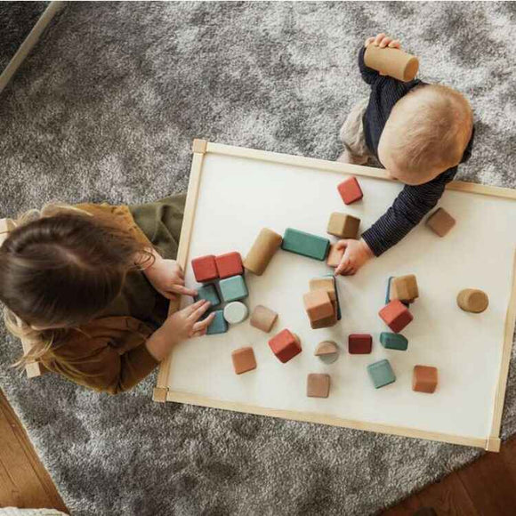 Two children play with the vibrant Korko Tall Architects, 40 Cork Building Blocks on a white table. One is light-haired, the other dark-haired, as they sit on a gray textured carpet, engaging in playful and educational activities that enhance fine motor skills through imaginative interaction.