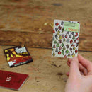 A hand holds a game card from The Kringle Caper by Big Potato, showcasing vibrant images and text, ideal for family game night fun. Nearby, additional cards are spread across a wooden surface. One prominently features the phrase "SECRET SANTA," while another displays the number "16," suggesting an exciting escape room adventure.