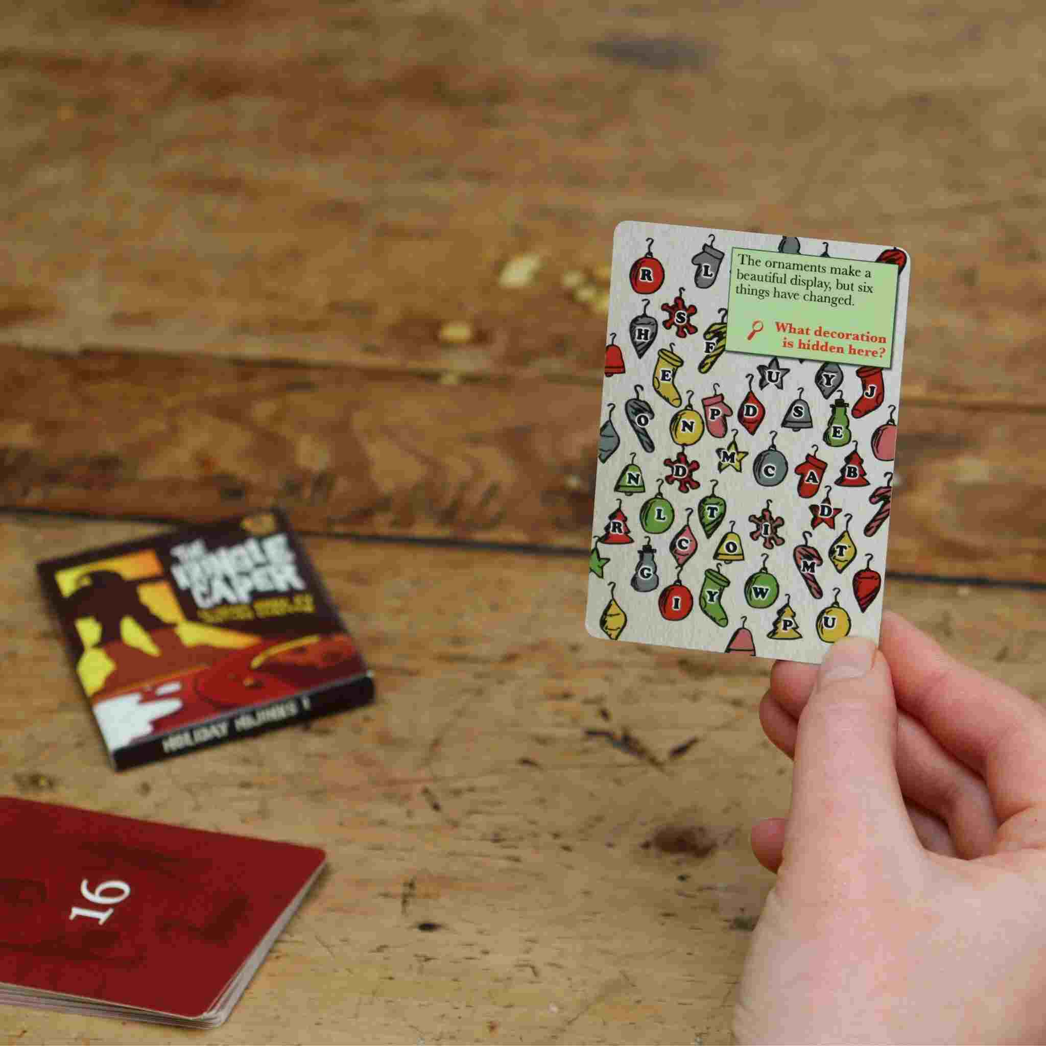 A hand holds a game card from The Kringle Caper by Big Potato, showcasing vibrant images and text, ideal for family game night fun. Nearby, additional cards are spread across a wooden surface. One prominently features the phrase "SECRET SANTA," while another displays the number "16," suggesting an exciting escape room adventure.