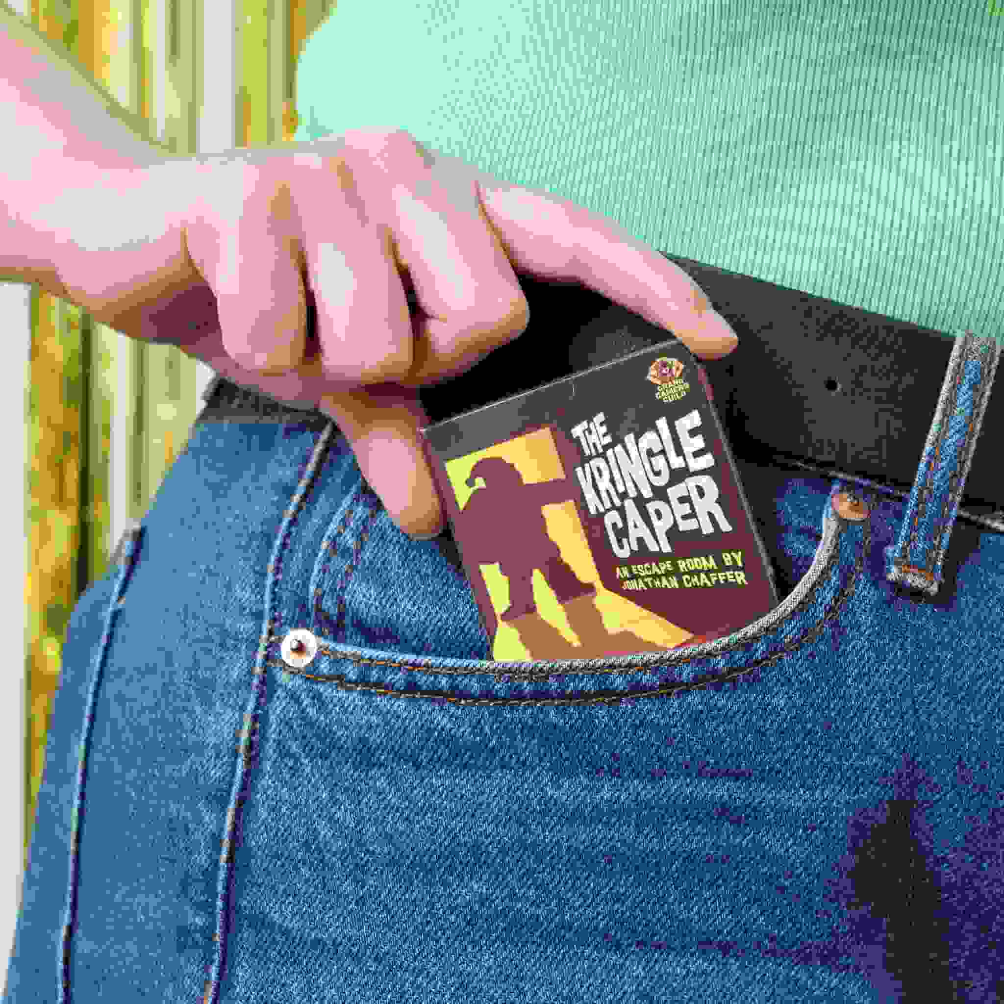 A person wearing jeans tucks a compact book titled *The Kringle Caper*, which resembles an escape room game, into their front pocket. The cover displays a silhouette set against a vibrant yellow and orange background. They are dressed in a green top, encapsulating the mystery-packed adventure crafted by Big Potato.